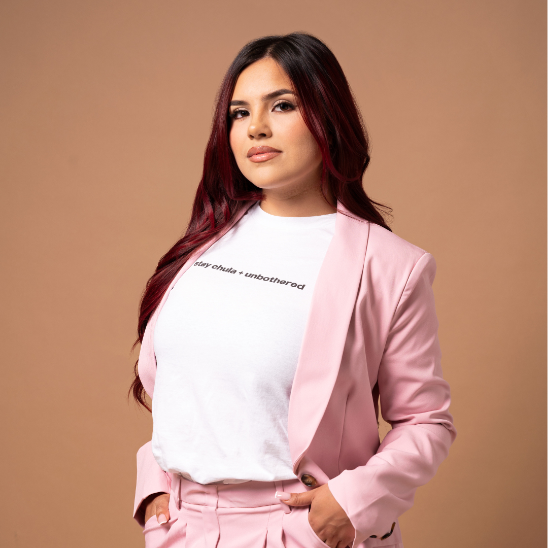 A woman with long, dark hair wears the Chula Baby Tee and Blazer & High Waisted Pants Set. The light pink blazer and pants are styled over a white t-shirt that reads "stay chula + unbothered" in black text. They pose against a beige background, looking directly into the camera with a composed expression.